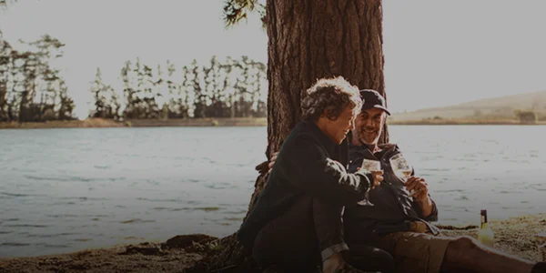 Couple sitting against tree trunk drinking wine with a lake in the background and tent beside them in the foreground